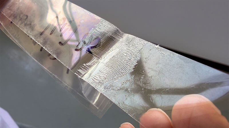 Close-up of a transparent release liner being peeled from an adhesive sample, showing visible residue or transfer marks after liner removal.